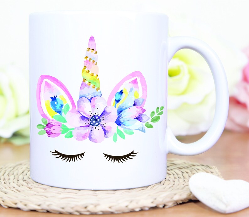 May include: White ceramic mug with a watercolor unicorn design. The unicorn has a pink and yellow mane, a gold horn, and closed eyes. The unicorn is surrounded by pink, purple, and blue flowers.