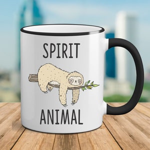 May include: White ceramic mug with a black rim and handle. The mug features a cartoon sloth hanging from a branch with the text "SPIRIT ANIMAL" printed on the mug.