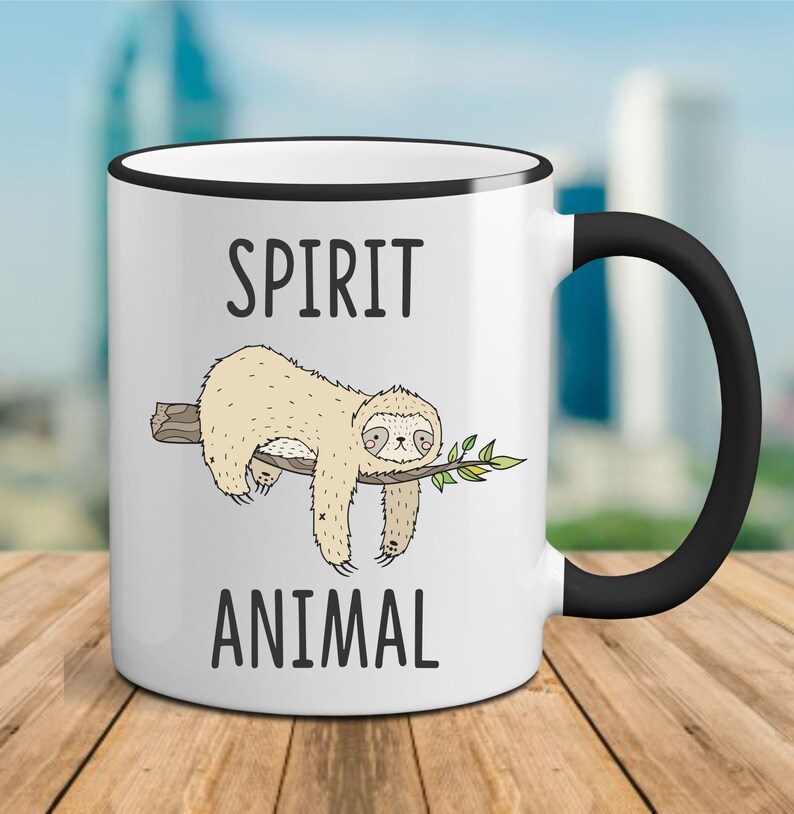 May include: White ceramic mug with a black rim and handle. The mug features a cartoon sloth hanging from a branch with the text "SPIRIT ANIMAL" printed on the mug.
