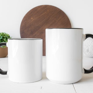 May include: Two white ceramic mugs with black handles and black rims. The mugs are on a white surface.