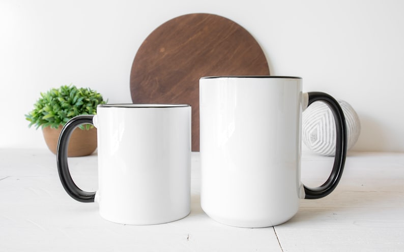 May include: Two white ceramic mugs with black handles and black rims. The mugs are on a white surface.