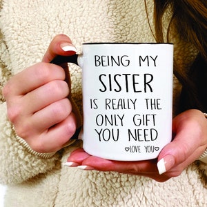 May include: White ceramic mug with a black handle and black text that reads "Being my sister is really the only gift you need. Love you."