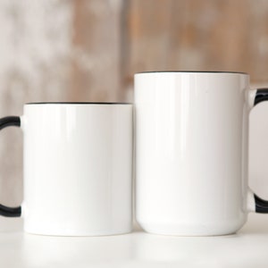 Op de afbeelding: Twee witte keramische mokken met zwarte handvatten en zwarte randen. De mokken zijn leeg en staan op een wit oppervlak.