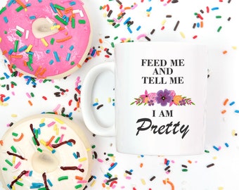 Könnte beinhalten: Weiße Keramiktasse mit schwarzem Text, der "Fütter mich und sag mir, dass ich hübsch bin" lautet, mit einem Blumendesign in Rosa, Lila und Gelb. Die Tasse ist von bunten Streuseln umgeben.