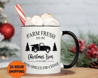 May include: White ceramic mug with a black handle. The mug features a black graphic of a pickup truck carrying a Christmas tree with the text "FARM FRESH EST. 1982 Christmas Trees CUT & CARRY PINE SPRUCE FIR CEDAR".