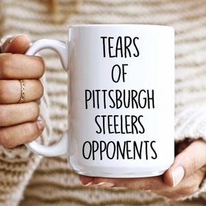 K&ouml;nnte beinhalten: Wei&szlig;e Keramik-Kaffeetasse mit schwarzem Text, der "Tears of Pittsburgh Steelers Opponents" lautet.