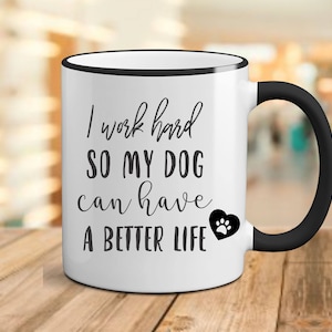May include: White ceramic coffee mug with a black rim and handle. The mug has black text that reads "I work hard so my dog can have a better life" with a black paw print heart.