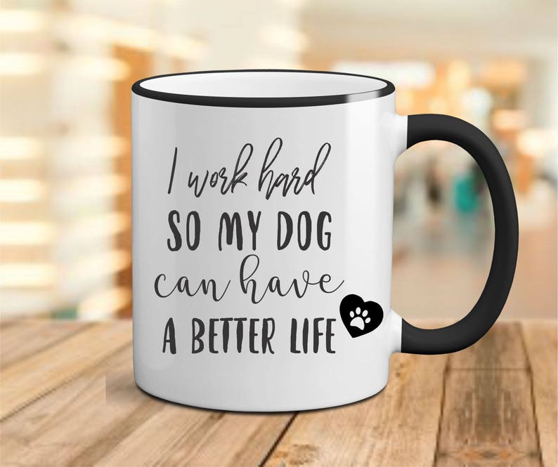May include: White ceramic coffee mug with a black rim and handle. The mug has black text that reads "I work hard so my dog can have a better life" with a black paw print heart.