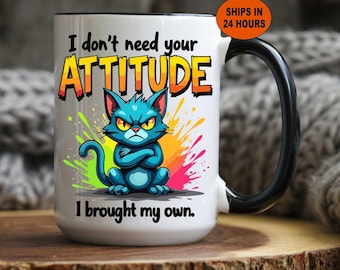 Könnte beinhalten: Weißer Keramikbecher mit schwarzem Griff und Rand, mit einer Cartoon-Katze in Blau mit wütendem Gesichtsausdruck und dem Text "I don't need your ATTITUDE, I brought my own." Bunte Farbspritzer umgeben die Katze.