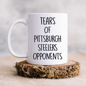 K&ouml;nnte beinhalten: Eine wei&szlig;e Keramik-Kaffeetasse mit schwarzem Text, der "Tears of Pittsburgh Steelers Opponents" lautet.