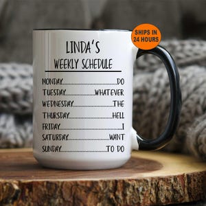 May include: White ceramic coffee mug with a black rim and handle. The mug has black text that reads "Linda's Weekly Schedule" and lists the days of the week with a fill-in-the-blank space for each day.