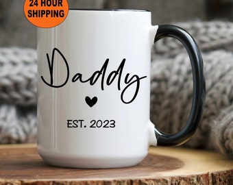 May include: White ceramic mug with a black rim and handle. The mug has black text that reads "Daddy" with a small heart and "EST. 2023" below.