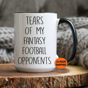 May include: White ceramic mug with black handle and rim. The mug features the text "TEARS OF MY FANTASY FOOTBALL OPPONENTS" in black, bold font. The mug is on a wooden surface.