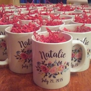 May include: A group of white ceramic mugs with floral designs and personalized names. Each mug has a different name, including "Natalie" and "July 21, 2018".