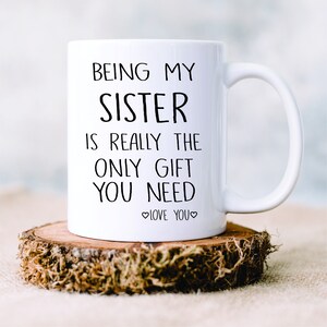 May include: White ceramic mug with black text that reads "Being my sister is really the only gift you need. Love you." The mug is sitting on a wooden slice.