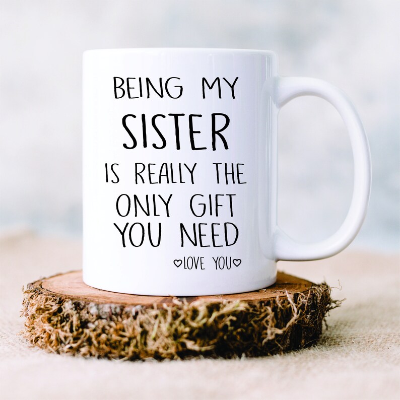 May include: White ceramic mug with black text that reads "Being my sister is really the only gift you need. Love you." The mug is sitting on a wooden slice.