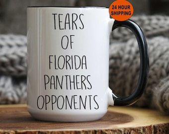 May include: White ceramic mug with a black handle and rim. The mug features the text "TEARS OF FLORIDA PANTHERS OPPONENTS" in black lettering. The mug is on a wooden surface.