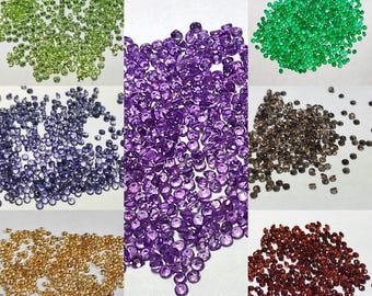 May include: Assortment of loose gemstones in various colors, including green, purple, brown, gold, and red. The gemstones are small and round, and they are scattered on a white surface.