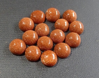 May include: A collection of polished, round, golden-brown gemstones with a sparkling, glittery appearance. The cabochons are arranged on a dark gray surface, showcasing their warm color and texture. These gemstones are ideal for jewelry making.
