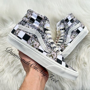 May include: White high-top sneakers with a black and white checkered pattern and floral print. The sneakers have a white sole and a row of rhinestones on the side.