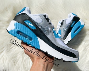 May include: White, gray, and black Nike Air Max 90 sneakers with blue accents and silver glitter on the swoosh logo. The sneakers have a white sole with a blue air bubble.
