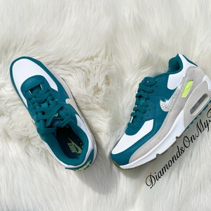 Puede incluir: Zapatillas Nike Air Max 90 blancas y verde azulado con un swoosh Nike plateado brillante y un acento amarillo en el lateral. Las zapatillas est&aacute;n sobre una superficie blanca y esponjosa.