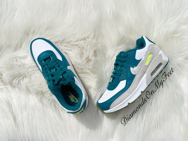 Puede incluir: Zapatillas Nike Air Max 90 blancas y verde azulado con un swoosh Nike plateado brillante y un acento amarillo en el lateral. Las zapatillas est&aacute;n sobre una superficie blanca y esponjosa.