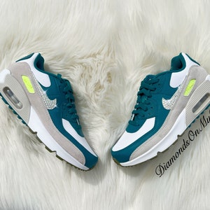 Puede incluir: Zapatillas de deporte de color azul verdoso y blanco con un swoosh blanco y diamantes de imitaci&oacute;n. Las zapatillas tienen una entresuela blanca y una suela gris. El texto "Diamonds On My Feet" es visible en la imagen.