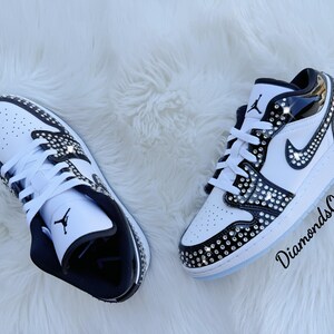 May include: Pair of white and black sneakers with crystal embellishments. The shoes feature a classic design with a perforated toe box, black accents, and a jeweled Nike swoosh. The soles are white with a light blue tint.