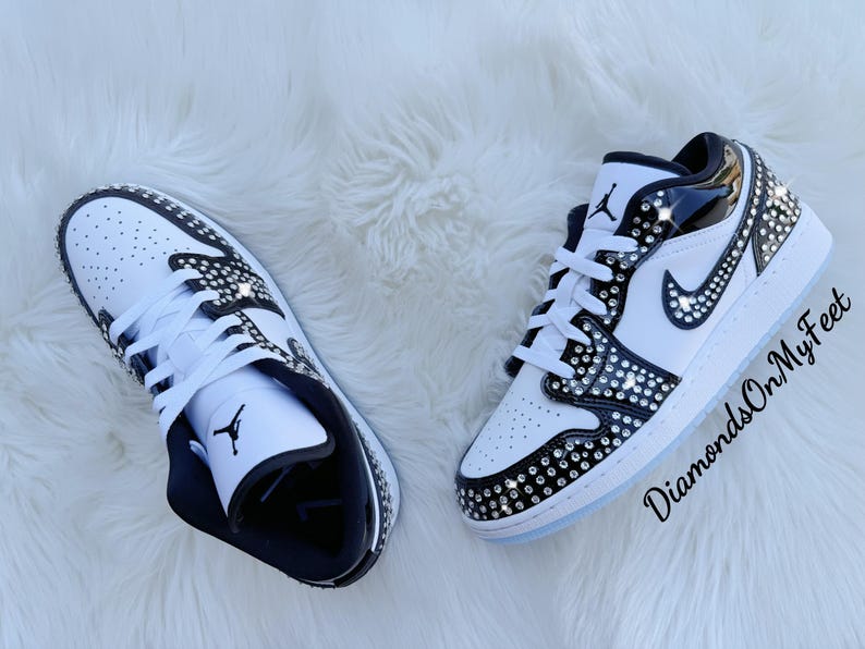 May include: Pair of white and black sneakers with crystal embellishments. The shoes feature a classic design with a perforated toe box, black accents, and a jeweled Nike swoosh. The soles are white with a light blue tint.