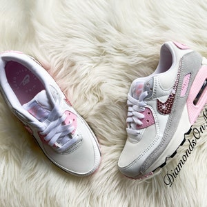 May include: White and pink Nike Air Max 90 sneakers with a pink swoosh and rhinestones. The sneakers have a white sole with a pink accent.