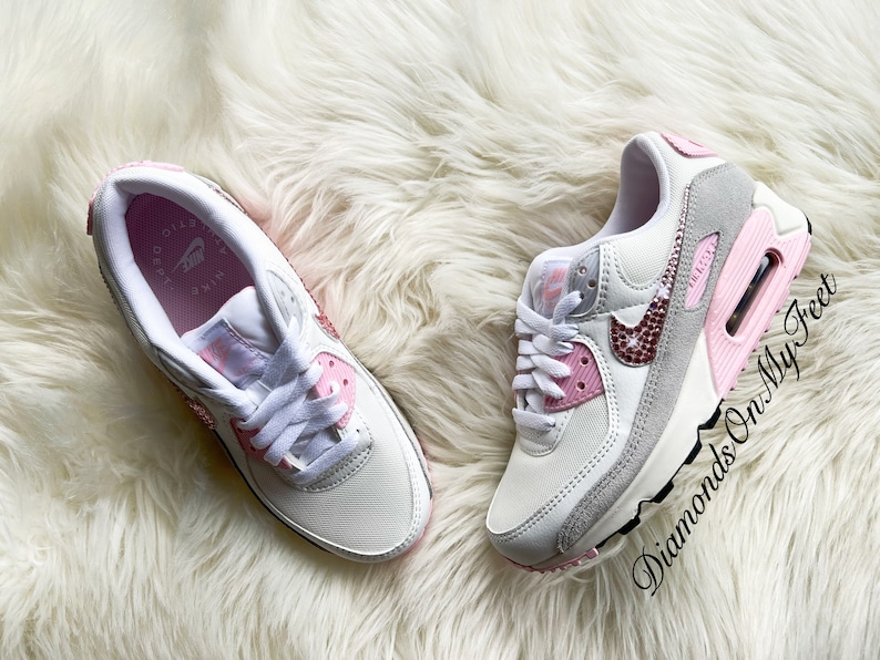 May include: White and pink Nike Air Max 90 sneakers with a pink swoosh and rhinestones. The sneakers have a white sole with a pink accent.