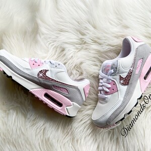 May include: A pair of white and grey Nike Air Max 90 sneakers with pink accents and rhinestones. The sneakers have a white swoosh logo and a pink and grey sole.
