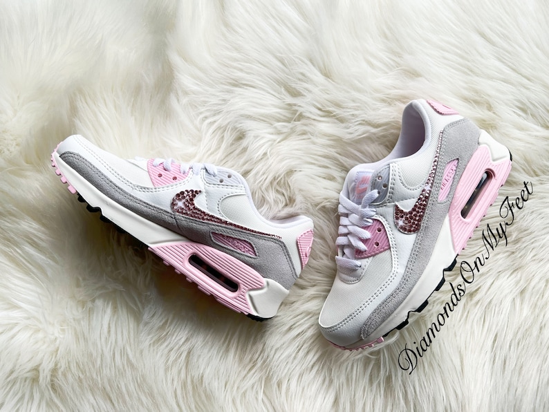 May include: A pair of white and grey Nike Air Max 90 sneakers with pink accents and rhinestones. The sneakers have a white swoosh logo and a pink and grey sole.