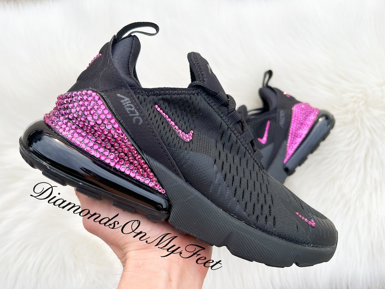 May include: Black Nike Air Max 270 sneakers with pink rhinestones on the heel and swoosh logo. The shoes are on a hand.