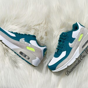 Puede incluir: Un par de zapatillas Nike Air Max 90 blancas y verde azulado con pedrer&iacute;a en el logotipo swoosh. Las zapatillas tienen una entresuela blanca y una suela gris. El texto "Diamonds On My Feet" es visible en la imagen.