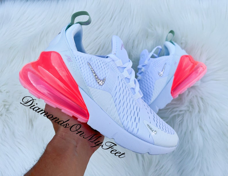 K&ouml;nnte beinhalten: Wei&szlig;e Sportschuhe mit Mesh-Obermaterial und einer leuchtend rosa-orangefarbenen Air-Bubble-Sohle. Der Nike-Swoosh ist mit Strasssteinen verziert. Der Text "Air 270" ist auf der Seite sichtbar.