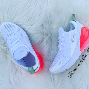 K&ouml;nnte beinhalten: Wei&szlig;e Nike Air Max 270 Sneaker mit einer leuchtend rosa Luftblase und Akzenten. Die Sneaker haben ein Kristall-Nike-Logo und liegen auf einer wei&szlig;en, flauschigen Oberfl&auml;che. Der Text "Diamonds On My Feet" ist sichtbar.
