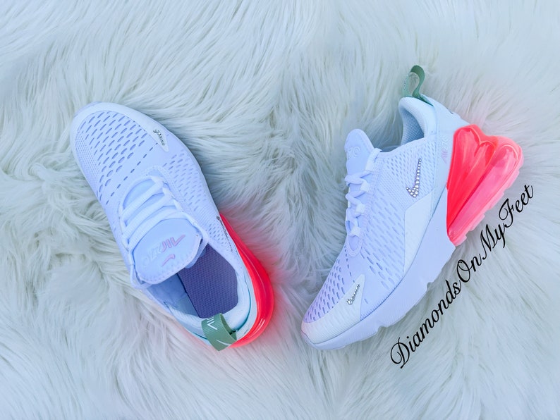 K&ouml;nnte beinhalten: Wei&szlig;e Nike Air Max 270 Sneaker mit einer leuchtend rosa Luftblase und Akzenten. Die Sneaker haben ein Kristall-Nike-Logo und liegen auf einer wei&szlig;en, flauschigen Oberfl&auml;che. Der Text "Diamonds On My Feet" ist sichtbar.