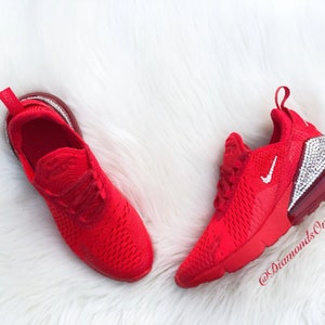 May include: Red Nike Air Max 270 sneakers with a white swoosh and a clear crystal-encrusted heel. The shoes are on a white fluffy surface. @DiamondsOnMyFeet
