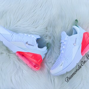 K&ouml;nnte beinhalten: Ein Paar wei&szlig;e Sportschuhe mit einem hellblauen Mesh-Obermaterial, einem Nike-Logo mit Schmucksteinen und einer leuchtend rosa-orangenen Luftblasen-Sohle. Die Schuhe liegen auf einer wei&szlig;en, flauschigen Oberfl&auml;che. Der Text "Diamonds On My Feet" ist kursiv geschrieben.