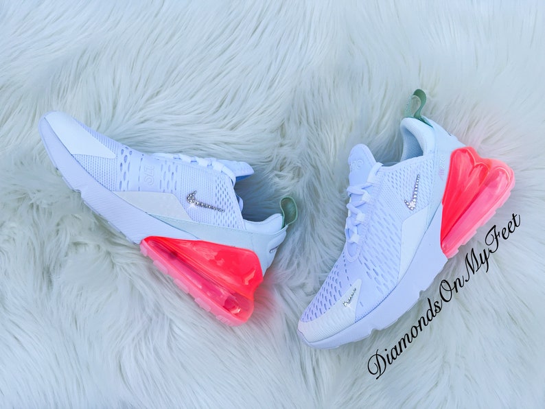 K&ouml;nnte beinhalten: Ein Paar wei&szlig;e Sportschuhe mit einem hellblauen Mesh-Obermaterial, einem Nike-Logo mit Schmucksteinen und einer leuchtend rosa-orangenen Luftblasen-Sohle. Die Schuhe liegen auf einer wei&szlig;en, flauschigen Oberfl&auml;che. Der Text "Diamonds On My Feet" ist kursiv geschrieben.