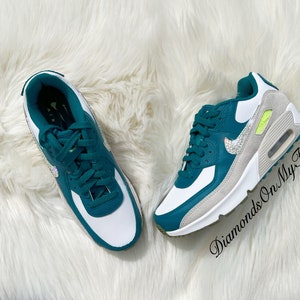 Puede incluir: Un par de zapatillas Nike Air Max 90 blancas y verde azulado con un swoosh blanco y diamantes de imitaci&oacute;n. Las zapatillas tienen una entresuela blanca y una suela exterior verde azulado. El texto "Diamonds On My Feet" es visible en la esquina inferior derecha de la imagen.