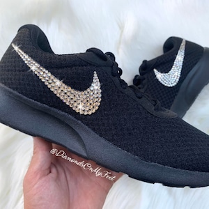 May include: Black Nike sneakers with a crystal-encrusted Swoosh logo. The shoes have a mesh upper and a white rubber sole.
