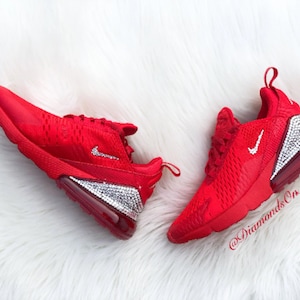May include: Red Nike Air Max 270 sneakers with a crystal-encrusted sole and a white Nike swoosh logo. The shoes are on a white fluffy surface. The text "@DiamondsOnMyFeet" is visible on the right shoe.