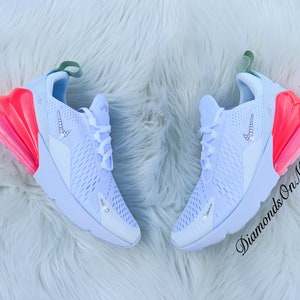 K&ouml;nnte beinhalten: Paar wei&szlig;e Sportschuhe mit einem hellblauen und rosa Farbverlauf. Die Schuhe haben ein kristallbesetztes Nike-Logo und Akzente. Der Text "Diamonds On My Feet" ist auf dem rechten Schuh sichtbar.