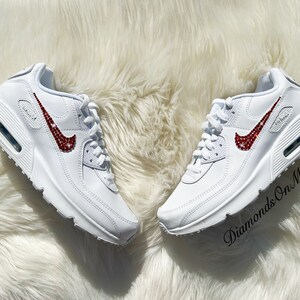 May include: White leather sneakers with a red rhinestone Nike swoosh. The shoes are on a white fluffy surface. The text "Diamonds On My Feet" is visible in the lower right corner of the image.