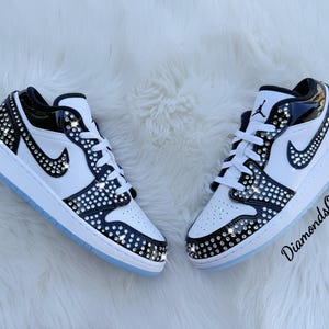 May include: Pair of white and black sneakers with crystal embellishments. The shoes feature a black swoosh, white laces, and a light blue sole. The sneakers are decorated with sparkling crystals.