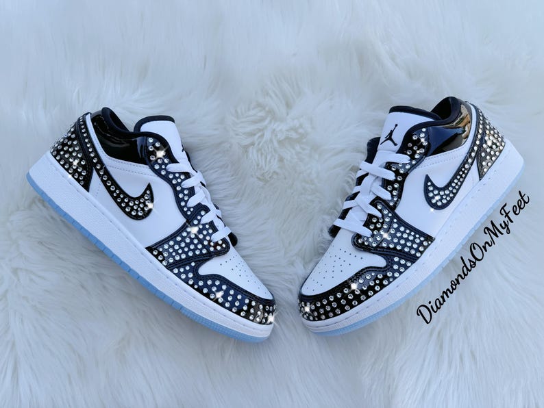 May include: Pair of white and black sneakers with crystal embellishments. The shoes feature a black swoosh, white laces, and a light blue sole. The sneakers are decorated with sparkling crystals.