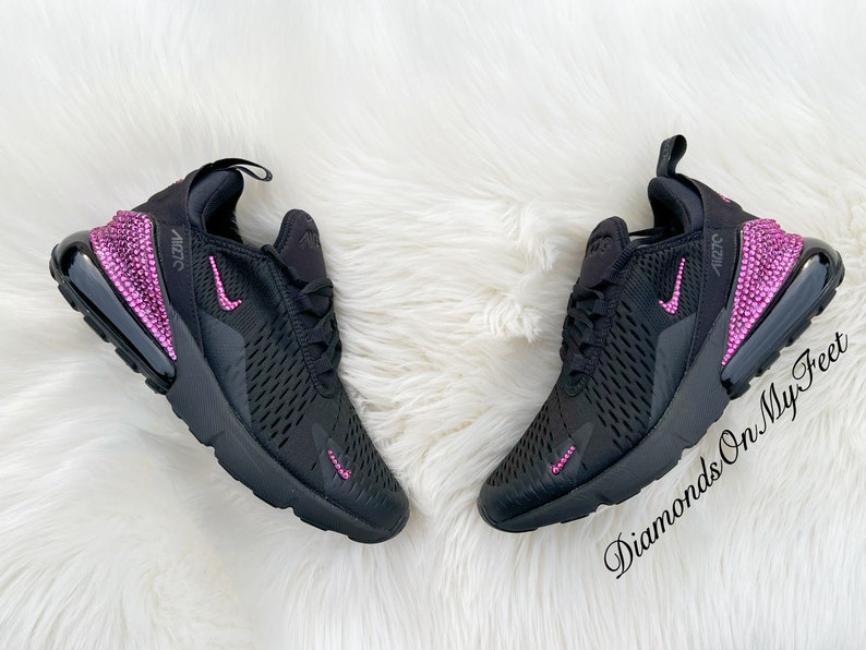 May include: Black Nike Air Max 270 sneakers with pink rhinestones on the heel and swoosh logo. The sneakers have a black mesh upper and a white fuzzy background.